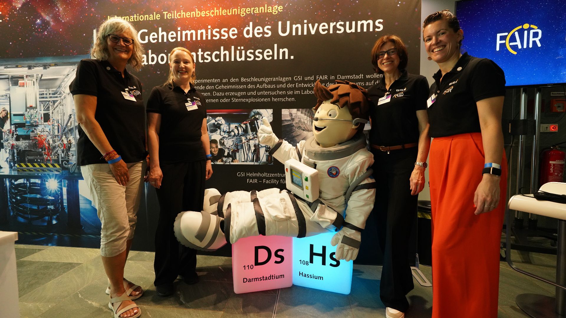 Das Team von GSI und FAIR am Expolink Messestand in Berlin zusammen mit einem Maskottchen auf leuchtenden Element-Würfeln.